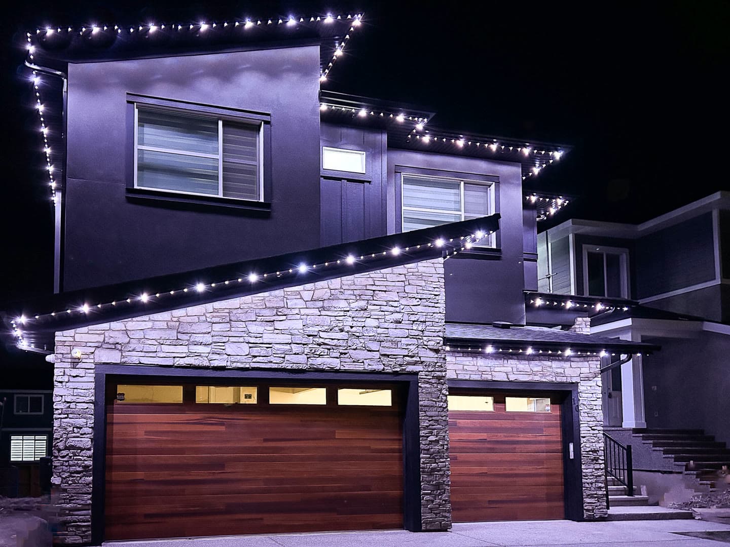 Modern farmhouse at dusk with warm-white permanent lights along the gables and porch